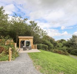A wooden pod with outdoor chairs on a patio surrounded by grass and trees at Woodland Pod Maerdy near Corwen