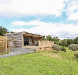 A wooden cabin with a deck and outdoor furniture on a grassy hill with trees in the background at Meadow Lodge Maerdy near Corwen