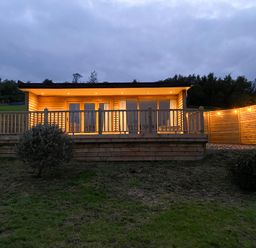 A wooden cabin with lights on a deck and a small tree in front at Meadow Lodge Maerdy near Corwen