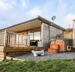 A wooden cabin with a deck railing a fire pit and a hot tub on a gravel area at Meadow Lodge Maerdy near Corwen