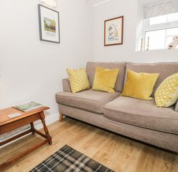 A living room with a beige sofa and yellow cushions next to a wooden side table with a lamp and magazines at Marlin Cottage in Conwy