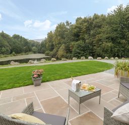 An outdoor patio with wicker chairs and a table with wine and grapes overlooking a grassy area with rocks and a pond surrounded by trees at Hendre in Penybontfawr