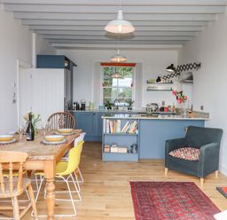 A kitchen and dining area with a wooden dining table set with bowls and glasses yellow chairs blue kitchen cabinets and a dark armchair with a patterned cushion at Wottonsbury in Ilsington