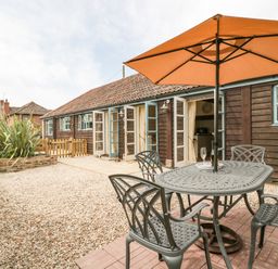 An outdoor patio area with a metal table and chairs under an umbrella in front of a wooden building with open double doors at Toms Barn Mudford Sock near Yeovil Somerset