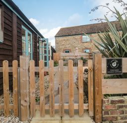 A wooden gate leading to a stone path between two buildings with blue windows at Tom's Barn in Mudford Sock near Yeovil Somerset