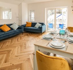 A dining area with a set table and a seating area with two blue sofas and orange and light blue cushions at Harbour Hideaway in Looe