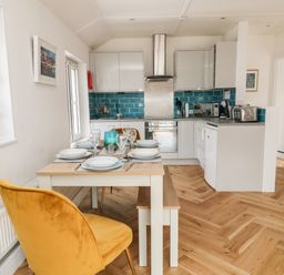 A kitchen with white cabinets a blue tile backsplash a dining table set for six with a yellow chair and wooden bench at Harbour Hideaway in Looe