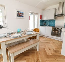 A kitchen and dining area with a wooden table set with plates glasses and cutlery alongside white cabinets and blue tiled backsplash at Harbour Hideaway in Looe
