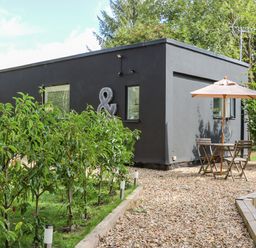 A dark gray modern house with small windows surrounded by greenery and a gravel patio with a wooden table and chairs under a beige umbrella at Kub House in Wigginton Hertfordshire