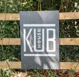 A gray sign with white text reading Kub House attached to a wooden fence with green foliage in the background at Kub House in Wigginton Hertfordshire