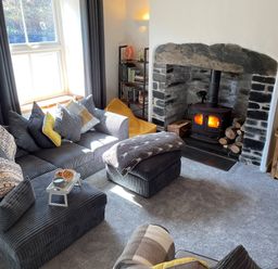 A living room with a gray sofa and cushions a stone fireplace with a wood stove and fire a television and wooden shelf at 7 Castle Terrace in Dolwyddelan