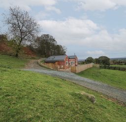 A small brick house with a fenced yard on a grassy hill with trees and a gravel driveway at Border View Lodge in Llandrinio