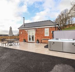 A small brick building with a patio area containing a table with four chairs and an umbrella and a hot tub next to a bench at Border View Lodge in Llandrinio