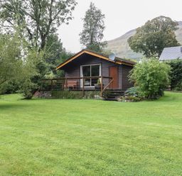 A small wooden cabin with a deck and a bench surrounded by grass and trees at Millmore Cabin in Killin