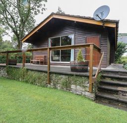 A wooden cabin with a patio area bench and potted plants and stairs leading to a grassy lawn at Millmore Cabin in Killin