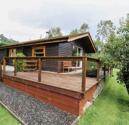 A wooden cabin with a raised deck and railing surrounded by grass and trees at Millmore Cabin in Killin