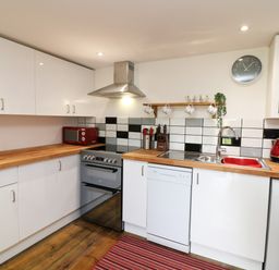 A kitchen with white cabinets wooden countertops a silver oven a red microwave a sink with a red basin a washing machine and utensils on a striped rug at Wisteria Cottage in Marldon