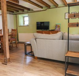 A living room with a gray sofa a wooden dining table with chairs a television on a wooden cabinet and exposed wooden beams on the ceiling at Bwthyn Gwyn Ty'n Y Pant in Boduan near Nefyn