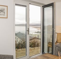 A living room with a wooden table a lamp on a side table gray sofas and glass doors overlooking a coastal view at The Sandy Bee in Benllech
