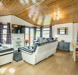 A living room with leather sofas a wall mounted tv a glass coffee table a dining table wooden ceiling and windows with curtains at Lakeview Lodge in South Lakeland Leisure Village