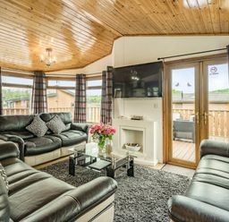 A living room with black leather sofas a glass coffee table a wall-mounted television and glass doors leading outside at Lakeview Lodge in South Lakeland Leisure Village