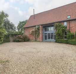 A gravel driveway leading to a brick house with a tiled roof and large windows surrounded by bushes and trees at The Barn in Tunstead near Hoveton & Wroxham