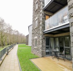An outdoor patio with table and chairs next to a stone building and a paved path with railing beside grassy areas at River View in Backbarrow