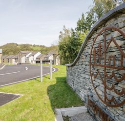 A stone wall with a metal house and tree design beside a curved road with houses and streetlights in the background at Ironworks in Backbarrow