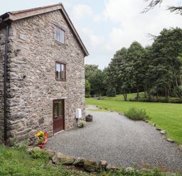 A stone building with small windows and a door surrounded by gravel and grass with trees in the background at Yr Hen Felin in Llanfyllin