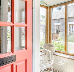 A porch area with a red door white chair and woven basket by windows at Sea Pickle Cottage in Llangwm Pembrokeshire