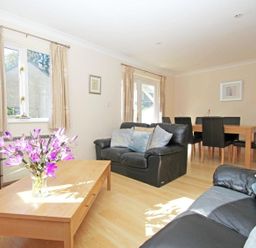 A living room with black leather sofas a wooden coffee table with purple flowers a dining table with black chairs and windows with beige curtains at Waters Edge in Falmouth