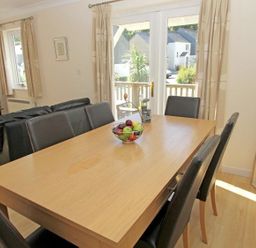 A dining area with a wooden table and six black chairs a fruit basket on the table a black leather sofa a television on a stand and glass doors leading outside at Waters Edge in Falmouth