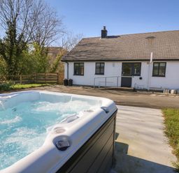 An outdoor hot tub with water jets in front of a white house with a sloped roof and fenced garden at Glyn Cottage in Llanon