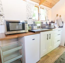 A kitchen area with a microwave on a wooden counter a small refrigerator a sink with a dish rack windows with patterned curtains and a fire extinguisher on the wall at Hugatree in Little Dunham near Litcham