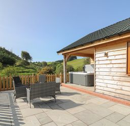 A wooden cabin patio with outdoor chairs and a hot tub overlooking green hills at Ploony Hill Cabin in Bleddfa near Knighton