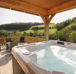 An outdoor patio with a hot tub and seating area overlooking green hills and trees at Ploony Hill Cabin in Bleddfa near Knighton