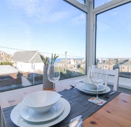 A dining table set with white bowls plates and wine glasses by a window overlooking rooftops and the sea at The Beach Hut in St Ives