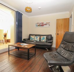 A living room with a black leather sofa and chair a wooden coffee table with coasters and a doorway with yellow chairs visible at Weybury Cottage Brewers Quay Harbour