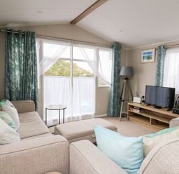 A living room with beige sofas cushions a small round table a TV on a wooden stand and blue patterned curtains at Ystwyth 26 in Borth