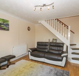 A living room with a leather sofa two stools a wall clock a radiator a staircase and a landscape picture at The Annex in Drybrook