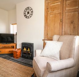 A living room with an armchair and a striped pillow next to a fireplace and a wooden cabinet at Hops Cottage in Doveridge