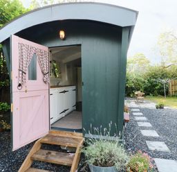 A green garden shed with a pink door open showing white cabinets inside a garden with a stone path and wooden table and chairs at Hazel Nook in Narberth