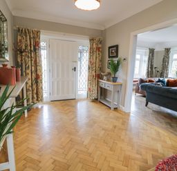 An entrance hall with wooden parquet flooring two tables with plants and decorative items a white front door and a view into a living room with sofas and windows at Pengwern in Llanrwst