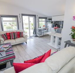A living room with two white sofas with red and striped cushions a TV on a striped bench a dining table with flowers and a kitchen area at The Garden House in Upper Quinton near Lower Quinton