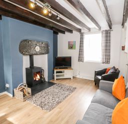 A living room with a grey sofa with orange cushions a black armchair a wood burning stove a television on a stand and exposed ceiling beams at Afallon in Penmachno