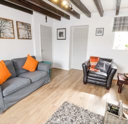 A living room with a gray sofa and orange pillows a black armchair with cushions a wooden side table with a lamp and bowl a window with plaid blind and framed pictures on white walls at Afallon in Penmachno