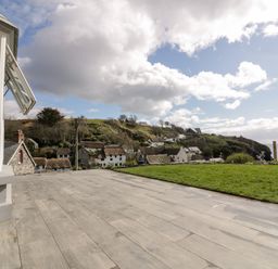 A patio with wooden flooring and a grassy area overlooking houses on a hillside by the sea at Louvain in Cadgwith near Ruan Minor