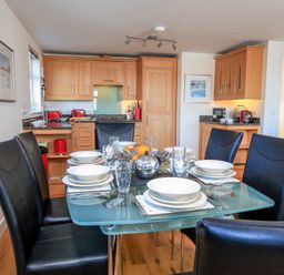 A dining area with a glass table set for six and black chairs in a kitchen with wooden cabinets at 2 The Bay in Bigbury