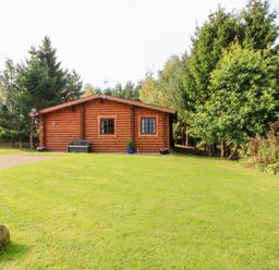 A log cabin with two windows a bench and potted plants on a grassy lawn surrounded by trees at Jalan End in Felton