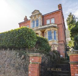 A red brick building with stone window frames behind a hedge and a stone wall with a gate reading St Heliers at The Hideaway in Malvern